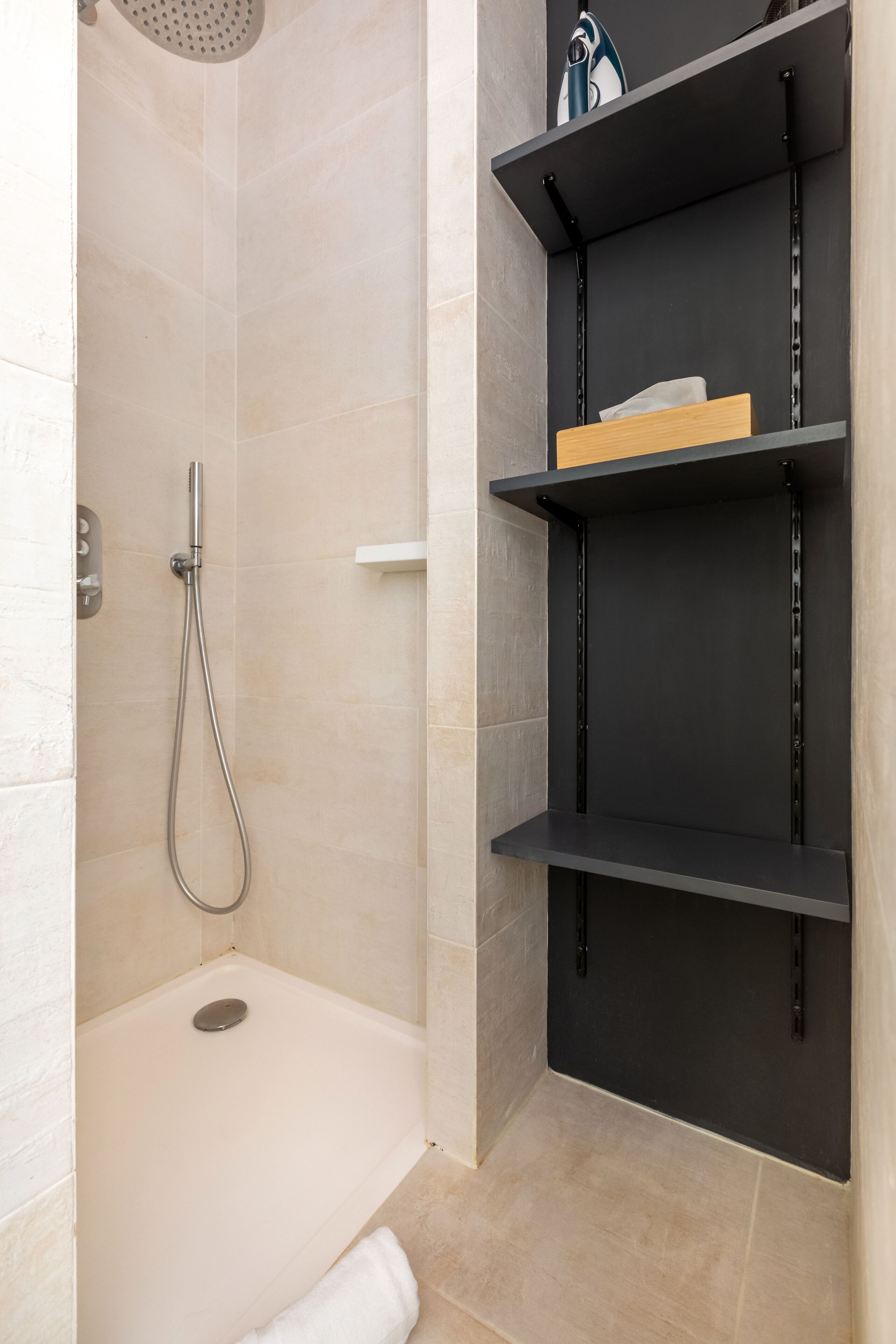 A shower with a handheld showerhead and a wall-mounted faucet, a white towel on the floor, and a black shelf unit with a box on the top shelf.