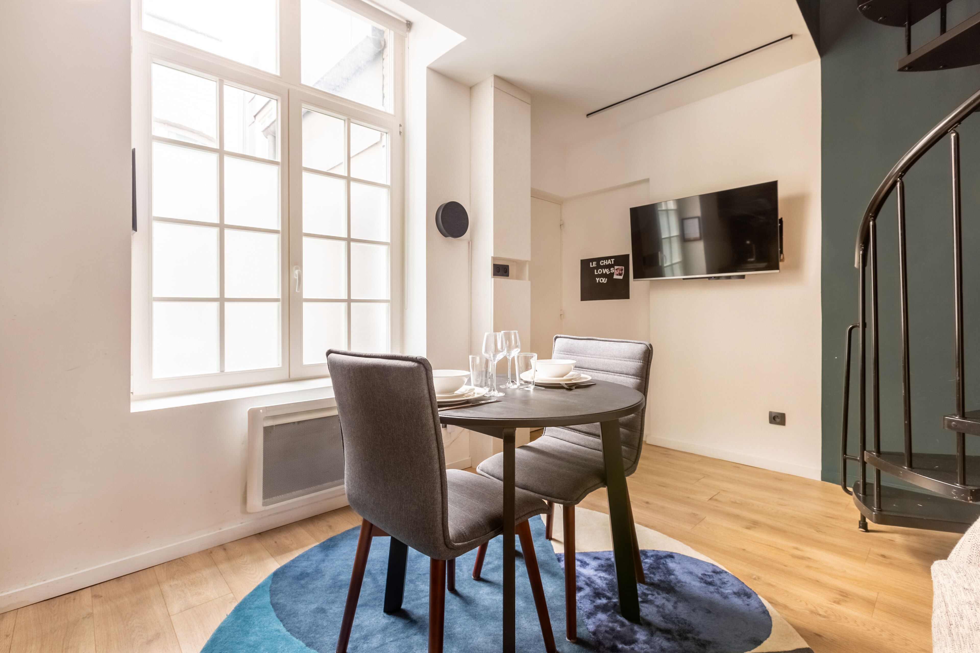 A dining area with a round table, two chairs, a window, a flat-screen TV, and a staircase.