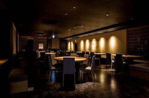 En restaurang med dämpad belysning, runda bord och stolar, en bardisk och en vägg med ett mönster som syns i bakgrunden.