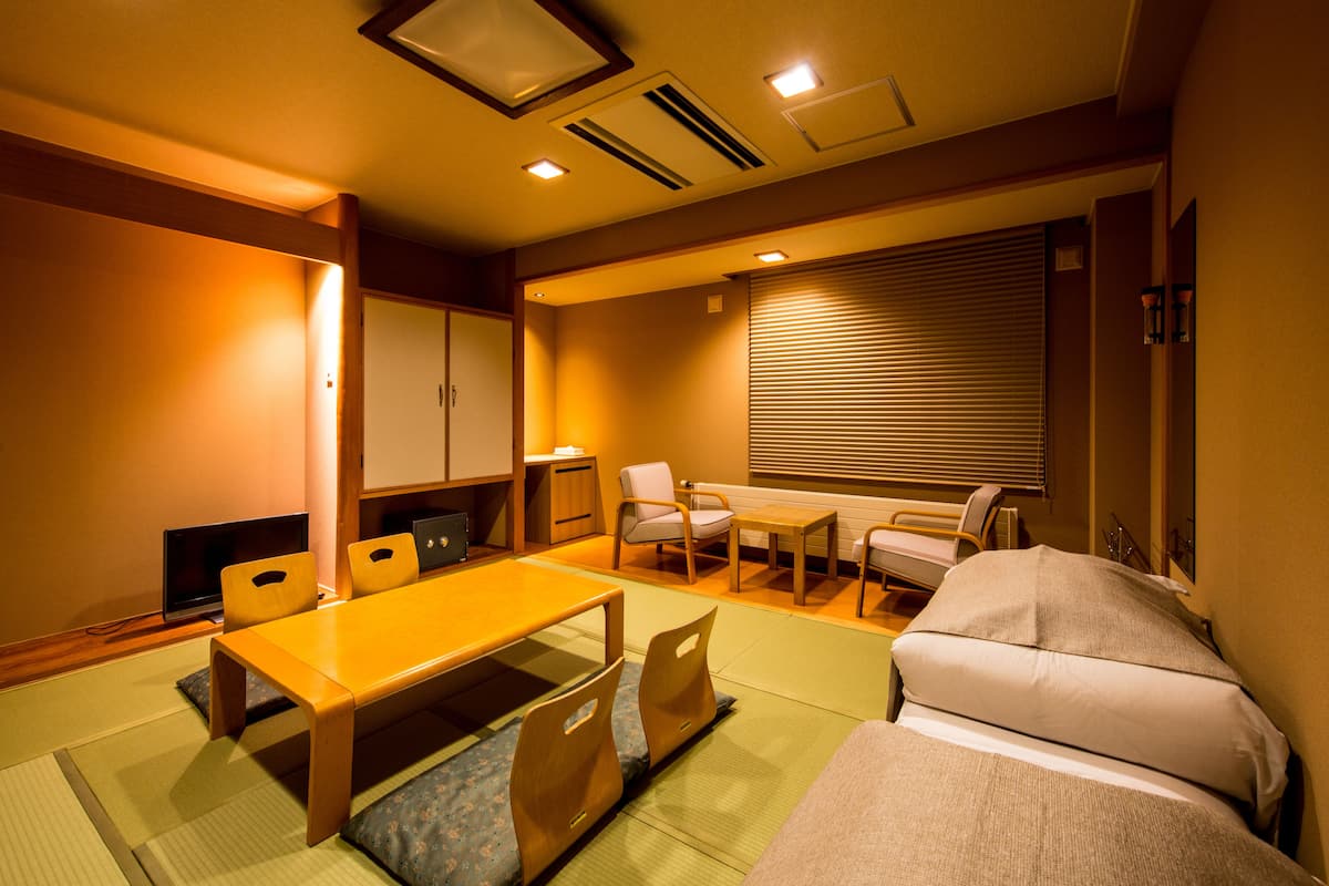 A hotel room with tatami mat flooring, a low wooden table, a sofa, a desk with a chair, and a sliding door with blinds.