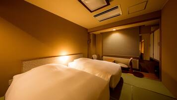 A hotel room with two beds, a bedside table, a chair, and a window with blinds.