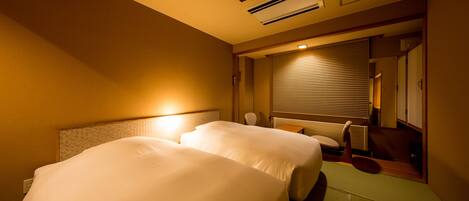 A hotel room with two beds, a bedside table, a chair, and a window with blinds.