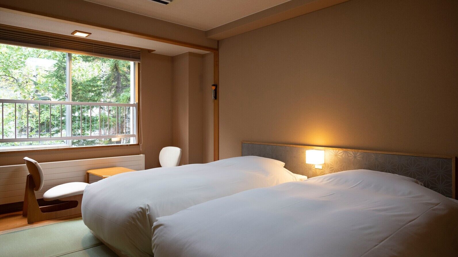 Quarto de hotel com duas camas, uma cadeira, uma mesa e uma janela com vista para árvores.
