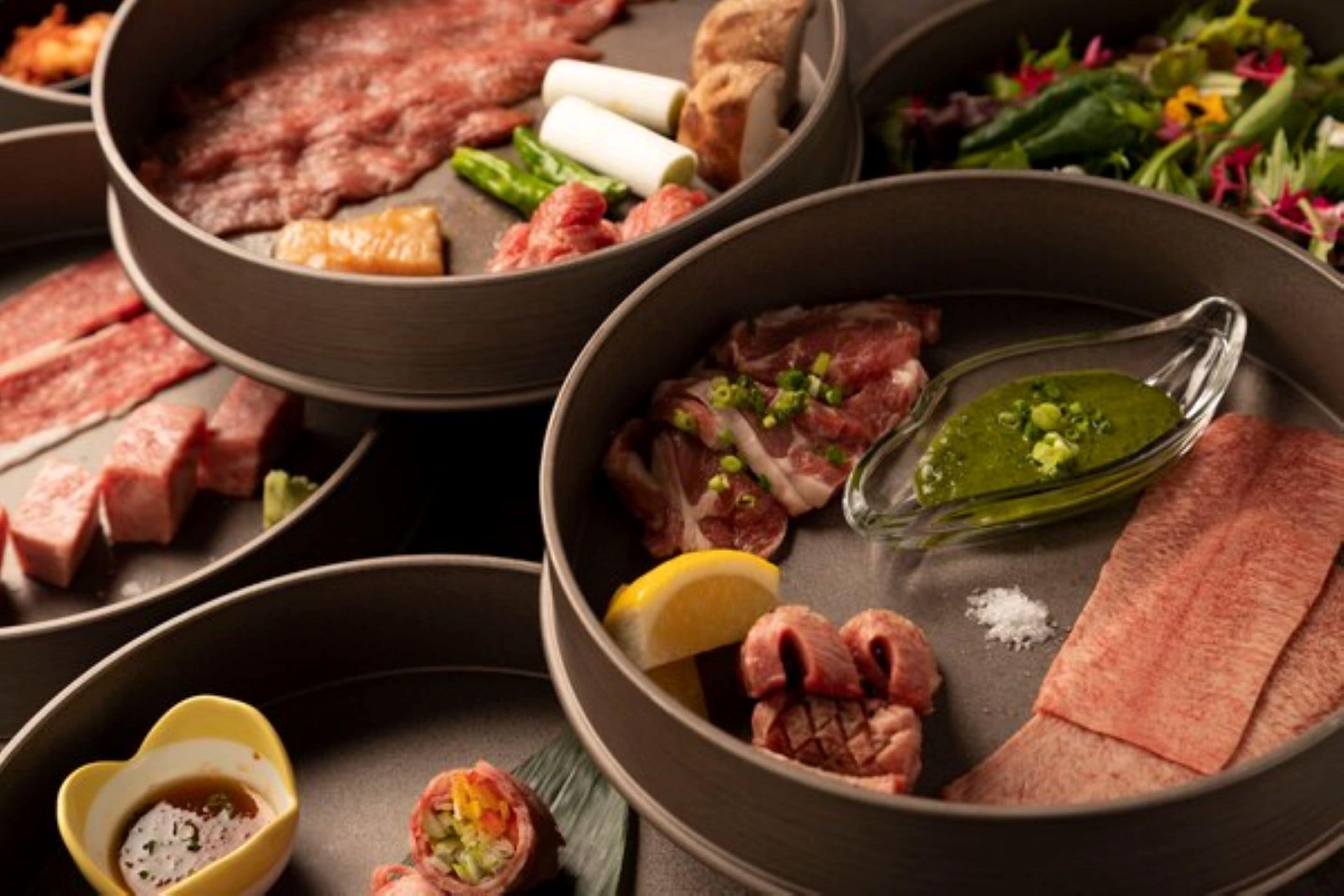 Variedad de carnes y verduras en una parrilla, con un recipiente pequeño de salsa y un envase con forma de corazón para condimentos.