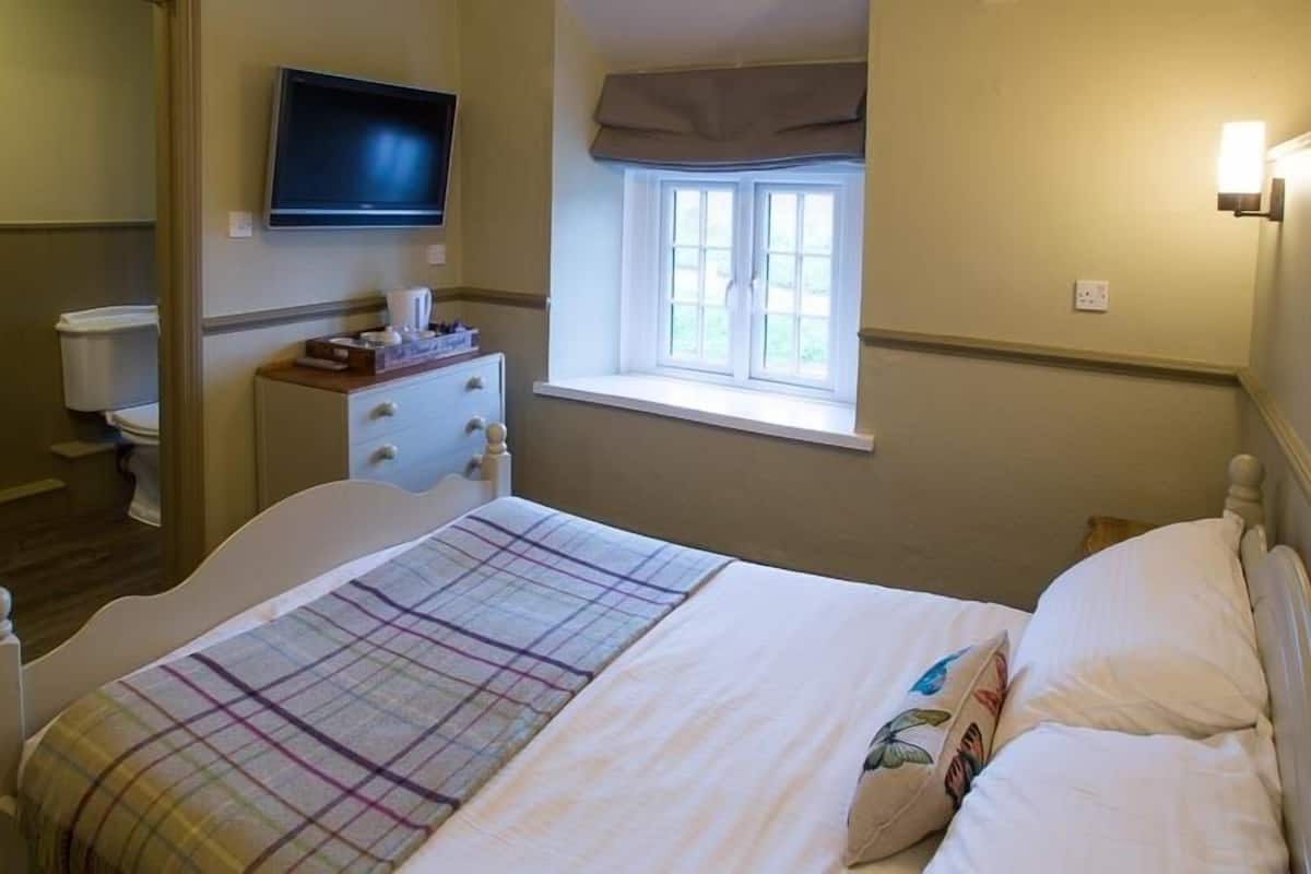 Un dormitorio con cama, televisión montada en la pared, ventana con vistas y mesita de noche con lámpara.