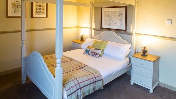 Un lit à baldaquin avec une couverture à carreaux, du linge de lit blanc et des coussins décoratifs. Il y a deux tables de chevet avec des lampes et une carte encadrée accrochée au mur.