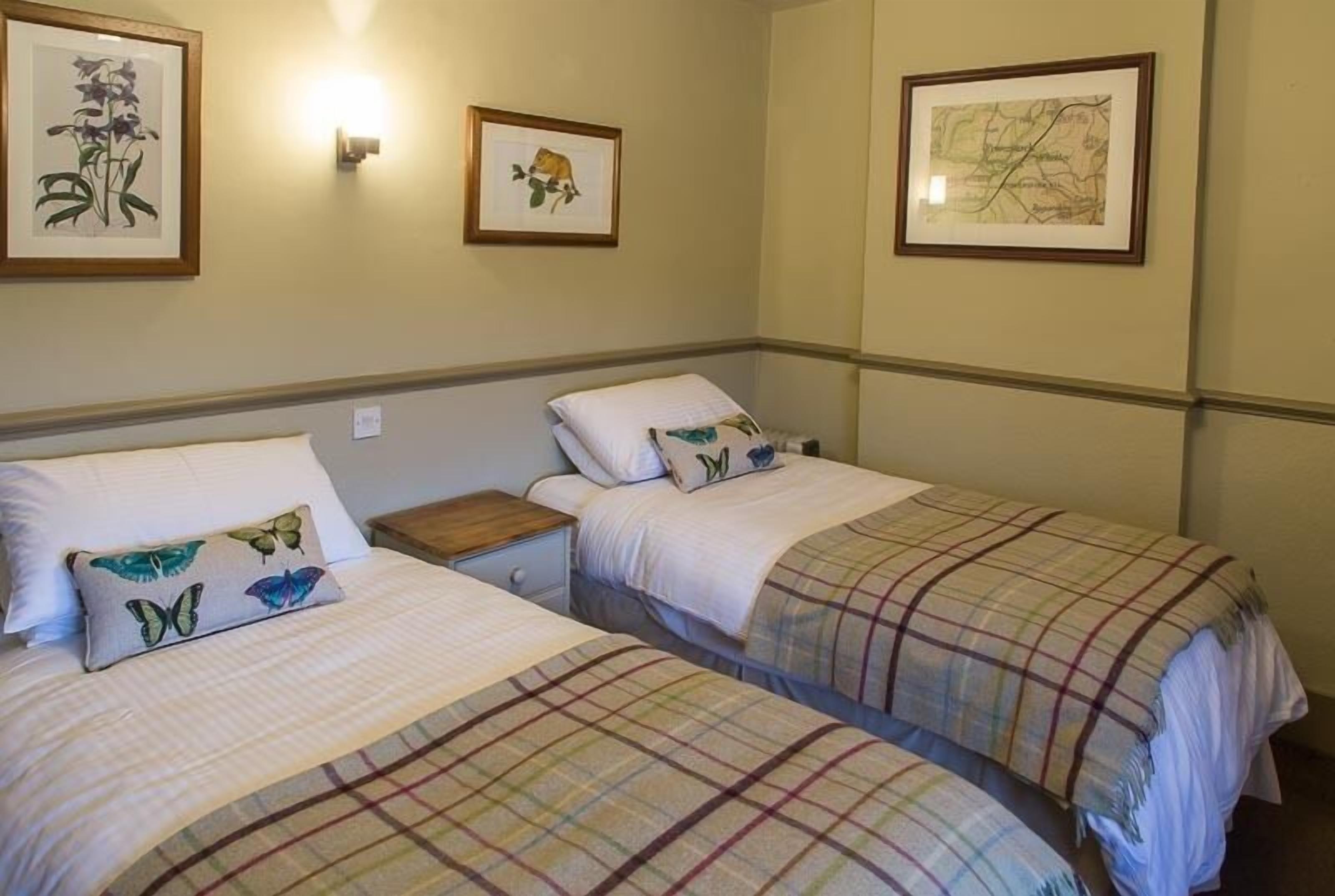 A hotel room with two beds, each with a plaid blanket and a pillow with a butterfly design. There are framed pictures on the wall, one of a plant and another of a map.