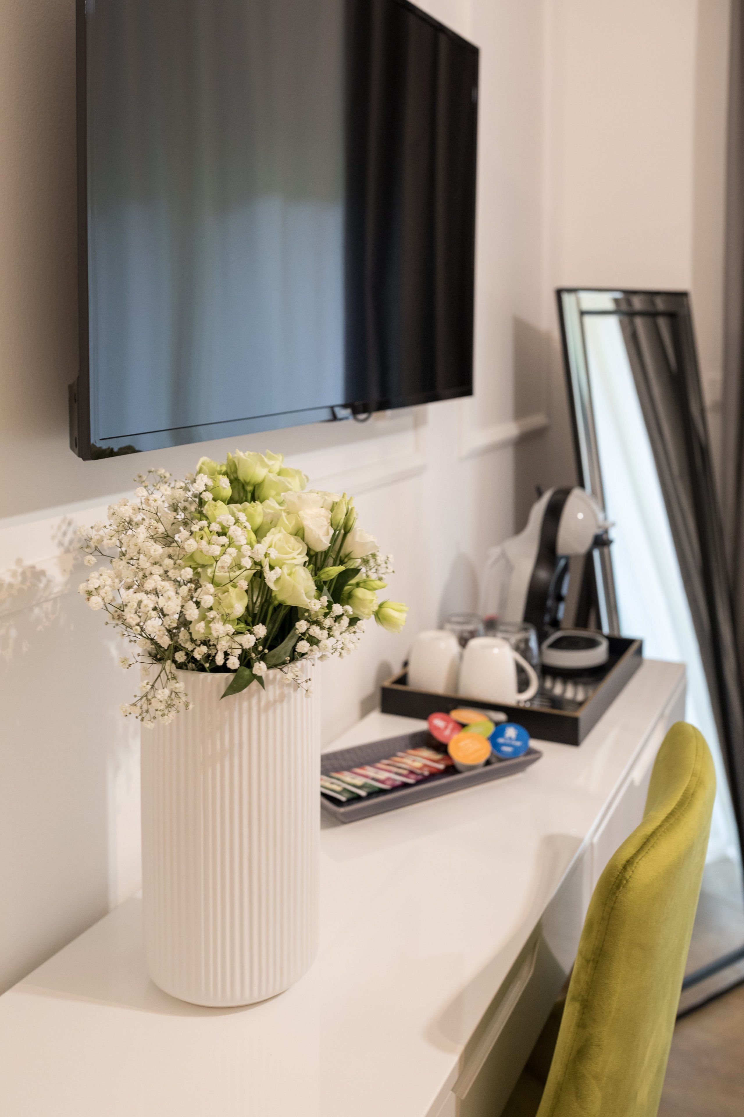 Un jarrón blanco con flores sobre una mesa blanca, un televisor negro montado en la pared, un espejo y una bandeja con tazas y un libro.
