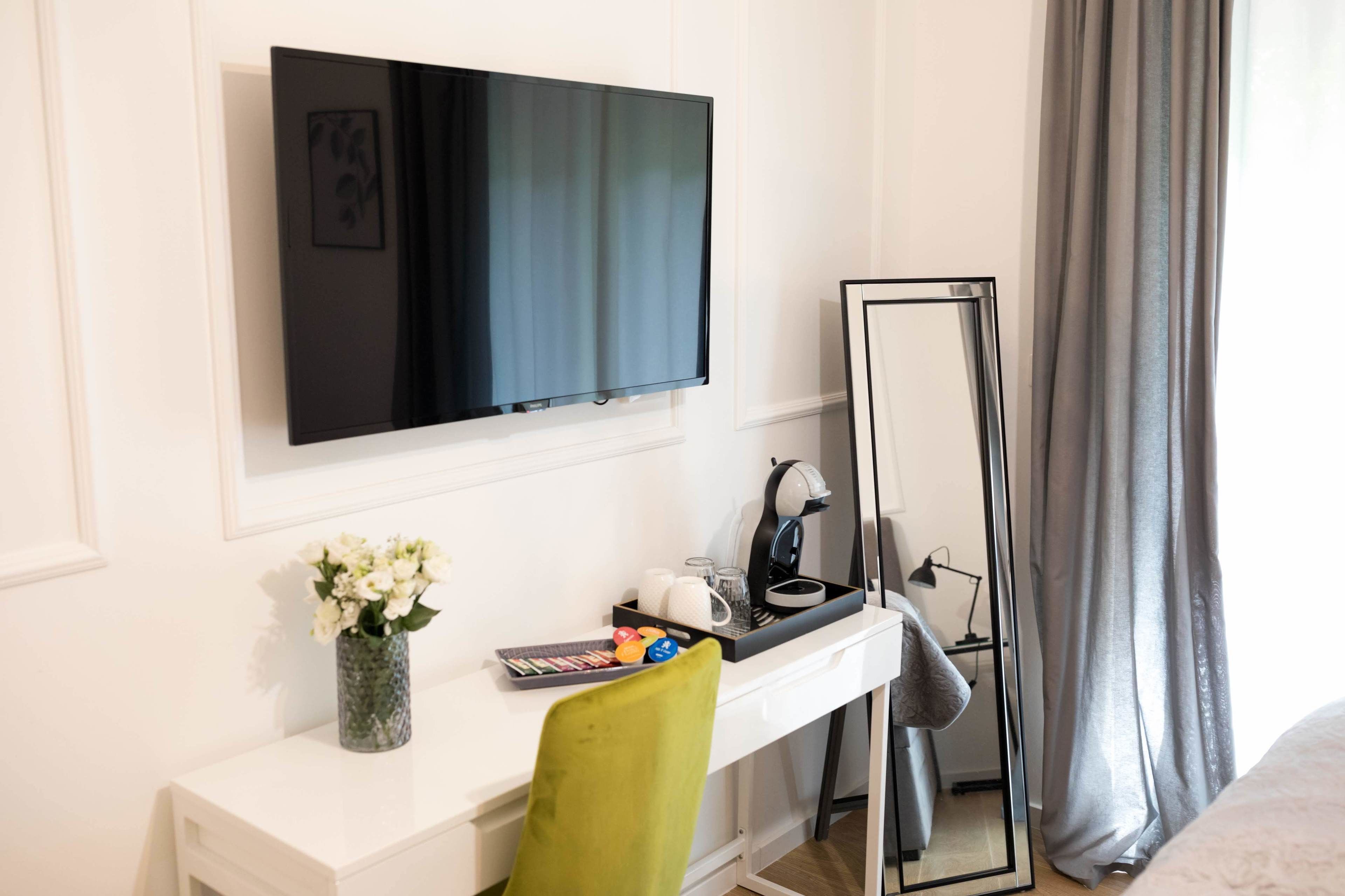 A hotel room with a TV mounted on the wall, a desk with a mirror, a vase of flowers, and a bed with a grey curtain.
