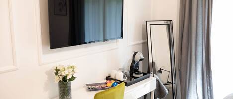 A hotel room with a TV mounted on the wall, a desk with a mirror, a vase of flowers, and a bed with a grey curtain.