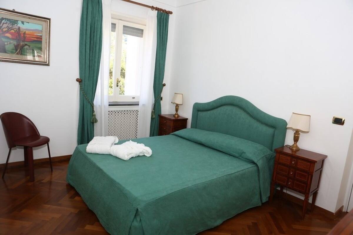 Un dormitorio con cama, una silla, una mesita de noche y una ventana con cortinas.