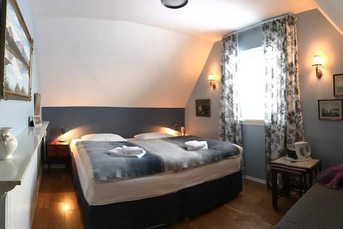 A bedroom with a bed, bedside tables, a nightstand, a window with curtains, and wall-mounted lights.