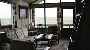 A living room with a sofa, armchair, and coffee table, a television mounted on the wall, and a view of the countryside through large windows. - Sel Guesthouse (Selfoss)