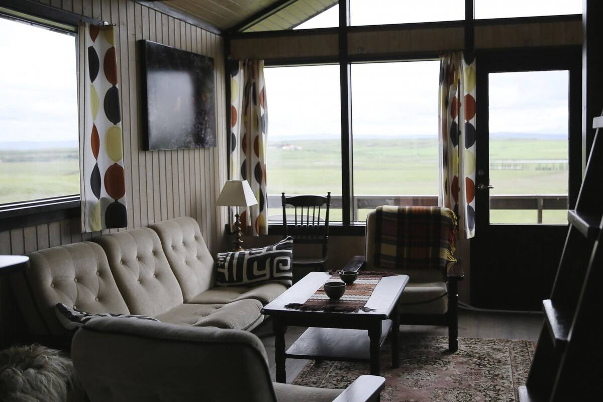 A living room with a sofa, armchair, and coffee table, a television mounted on the wall, and a view of the countryside through large windows.