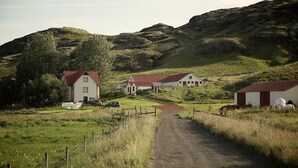 A rural landscape with a dirt road leading to a cluster of white buildings with red roofs, surrounded by green fields and hills. - Sel Guesthouse (Selfoss)