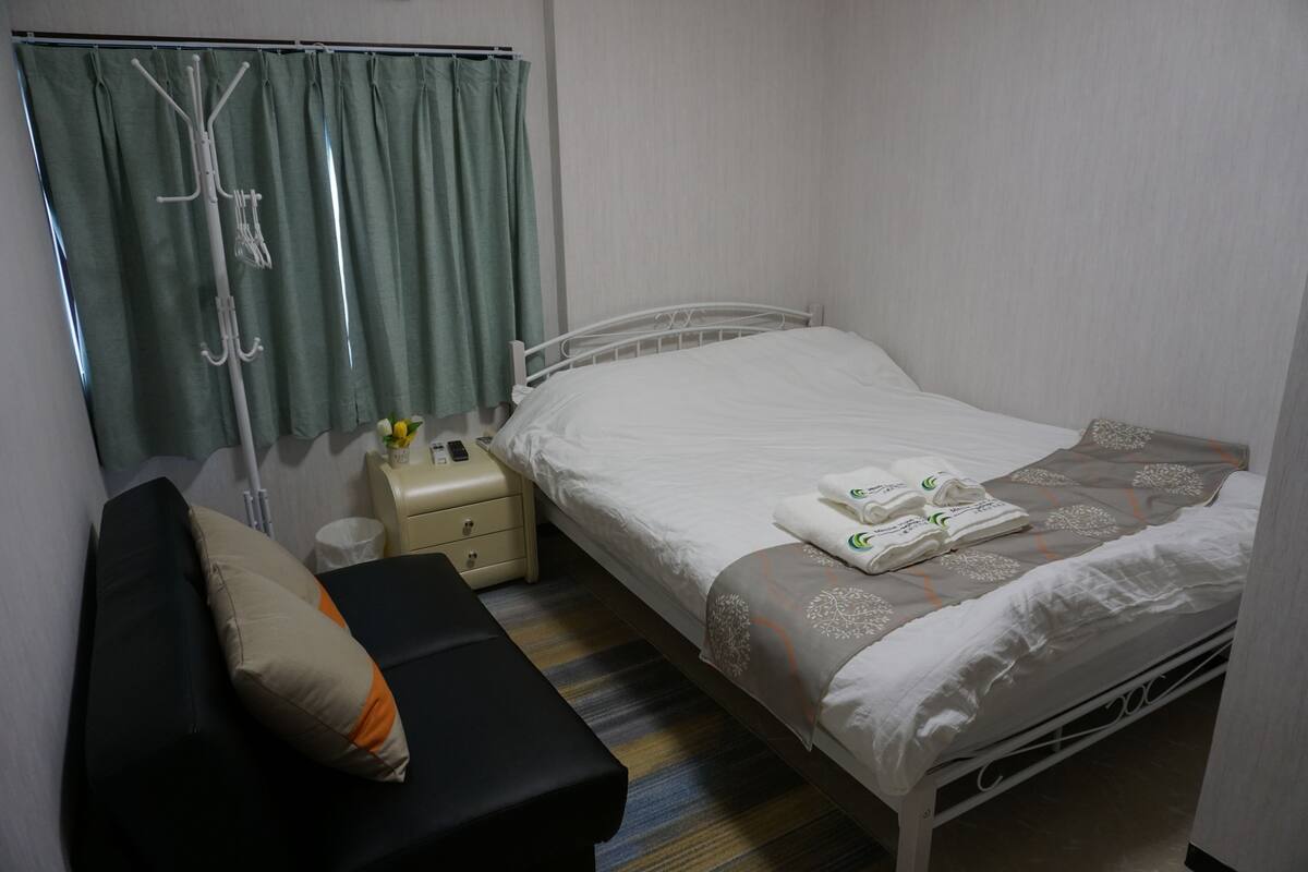 Una habitación de hotel con una cama, una mesita de noche, una lámpara, un banco y una cortina.