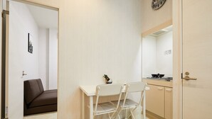 Habitación compacta con un escritorio blanco, dos sillas, una pequeña planta en maceta y una zona de cocina con estufa.