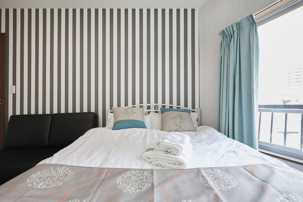 Una habitación de hotel con cama, un sofá negro, un pared a rayas y una ventana con cortinas.