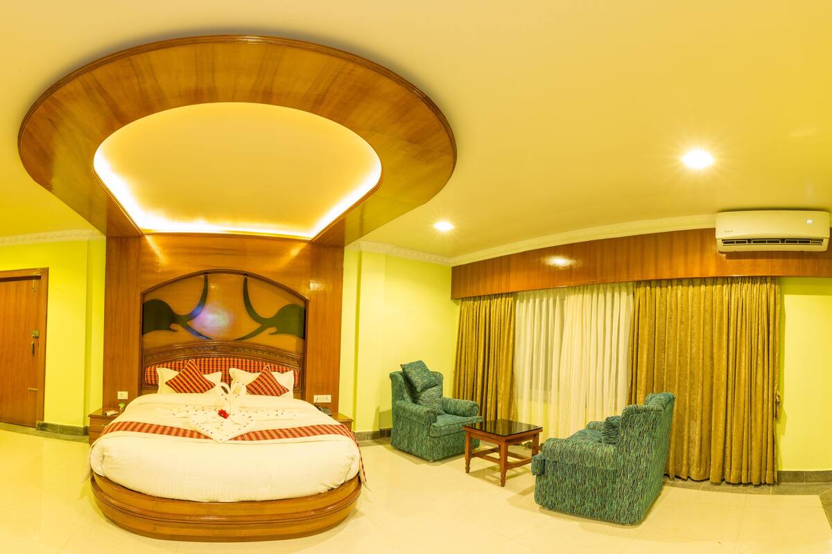 A hotel room with a large bed, a wooden headboard, a round ceiling light, a green armchair, and a small table.