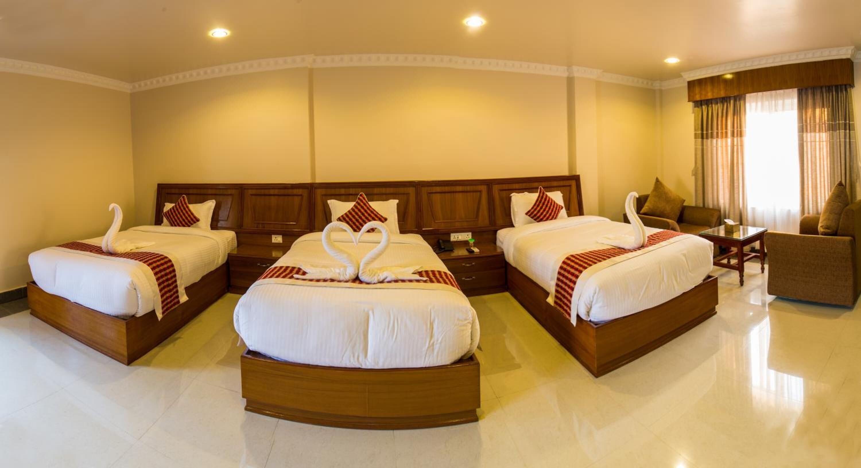 A hotel room with three beds, each with a wooden headboard and white bedding. There is a small table and a chair in the corner.