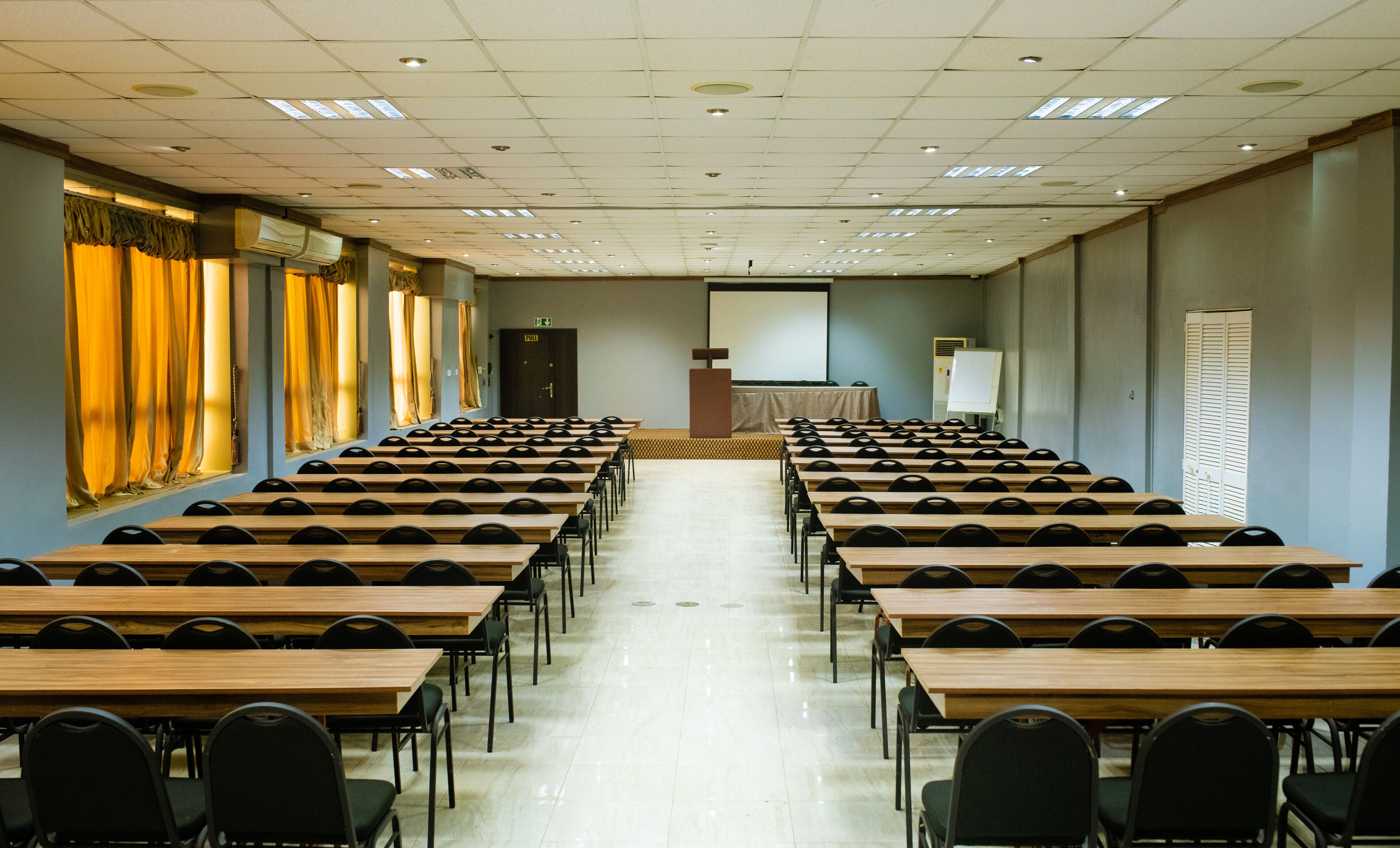 A conference room with rows of wooden tables and black chairs, a podium, and a projection screen.