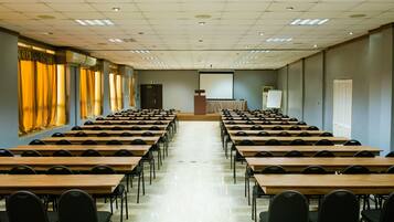 A conference room with rows of wooden tables and black chairs, a podium, and a projection screen.