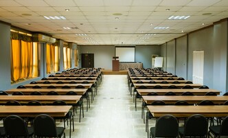 A conference room with rows of wooden tables and black chairs, a podium, and a projection screen.