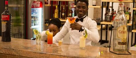 Un espace bar avec un comptoir, un homme préparant une boisson et, au fond, des étagères remplies de bouteilles.
