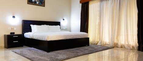 A bedroom with a large bed, a nightstand, a wall-mounted light, and a window with curtains.