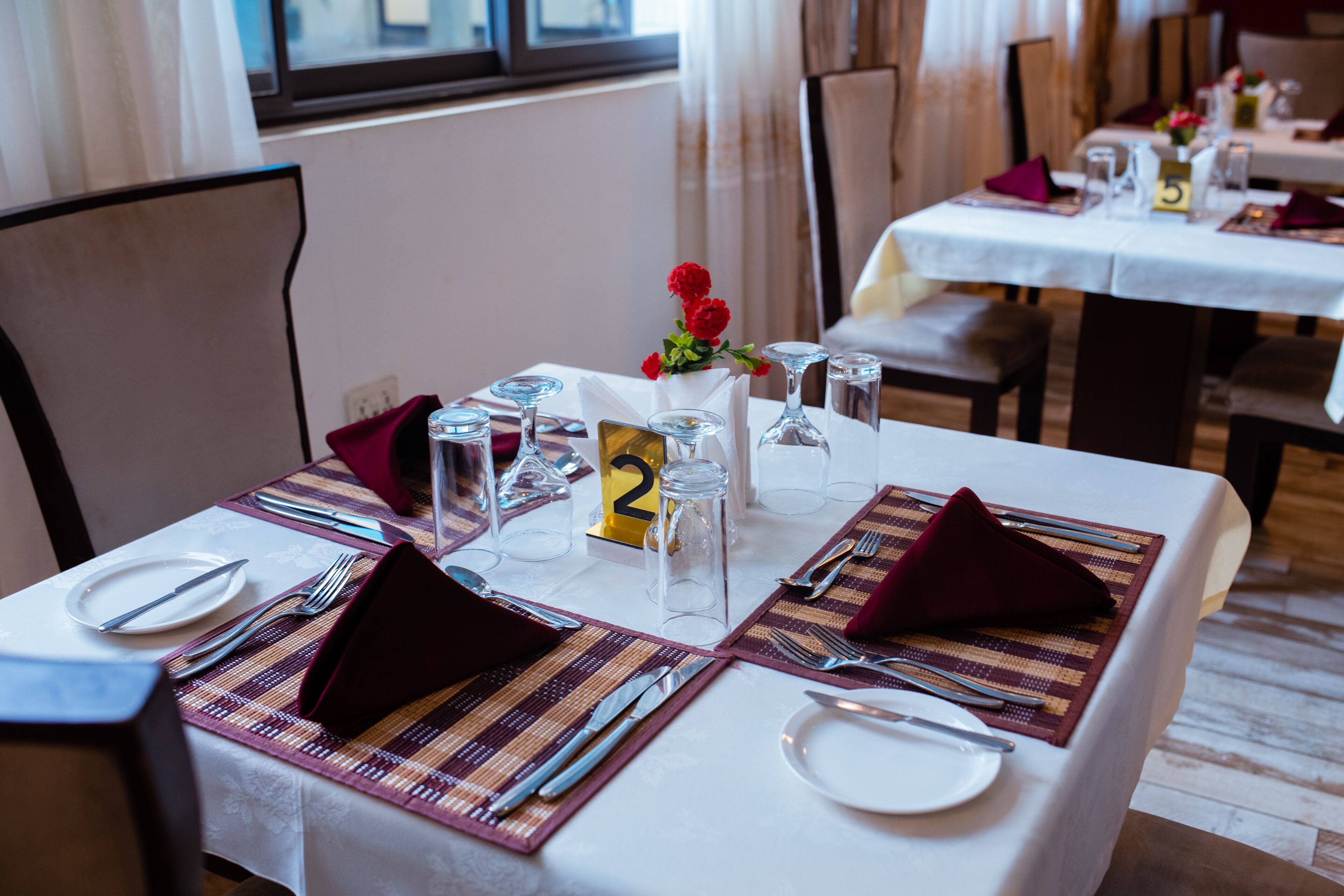 Una mesa de comedor dispuesta con puestos numerados, vasos transparentes y un pequeño jarrón con flores rojas.