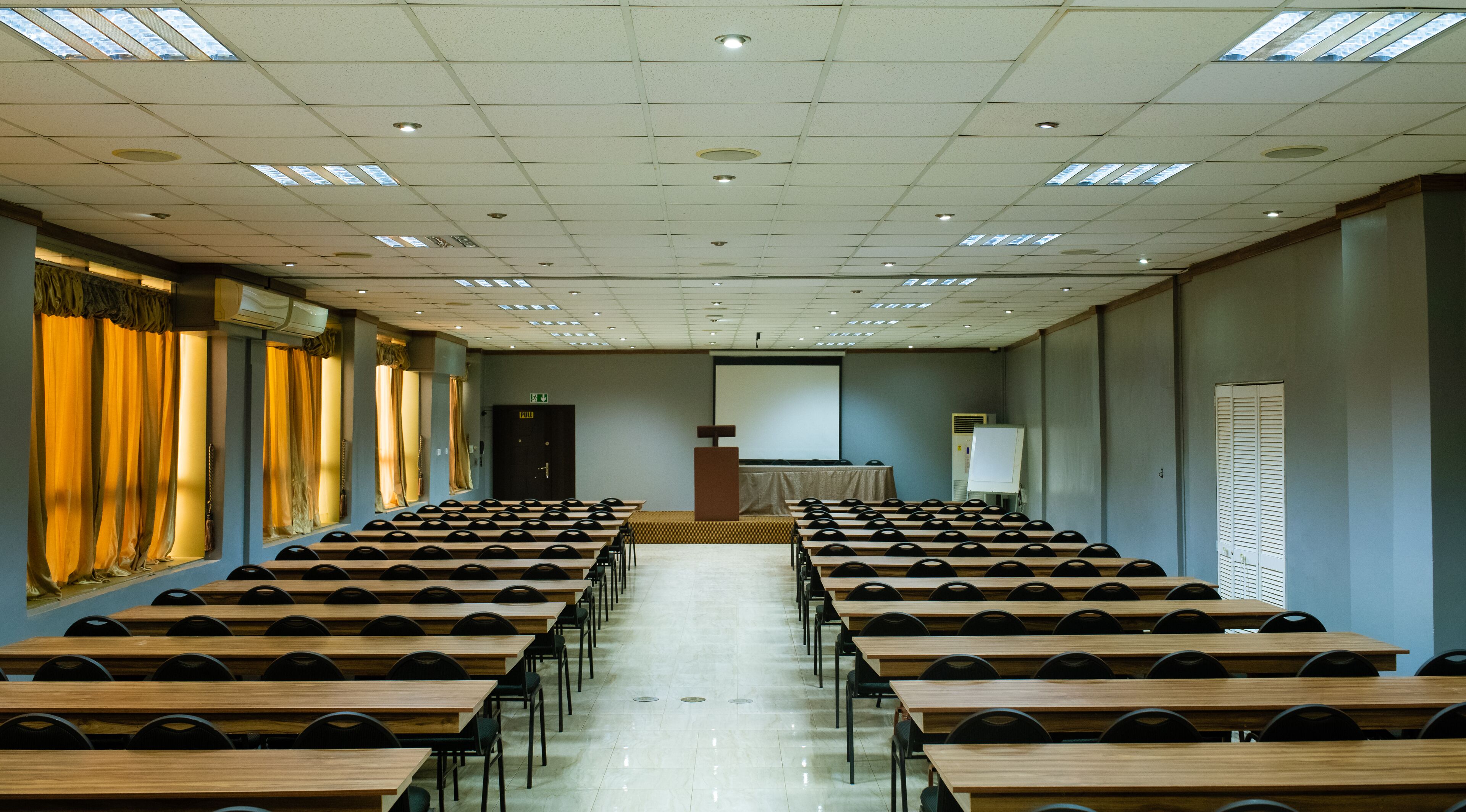 A conference room with rows of wooden tables and chairs, a podium, a projector screen, and a pull-down door.