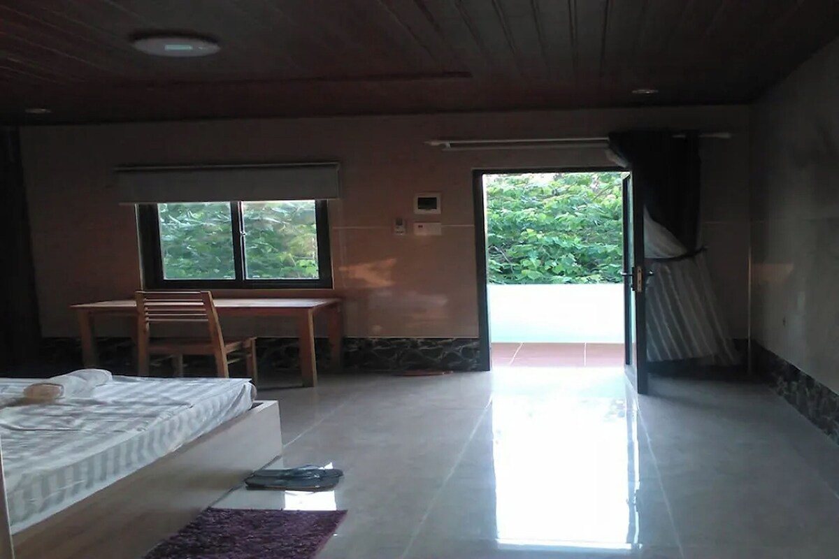 Ein Schlafzimmer mit Bett, Schreibtisch und Stuhl. Ein Fenster bietet Blick auf grüne Natur.