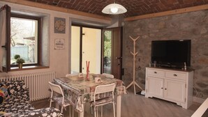A cozy living room with a brick ceiling, a dining table with chairs, a sofa, a television on a white cabinet, and a clock on the wall.