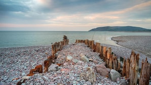 Une plage de galets bordée d’une clôture en bois, avec une montagne qui se profile à l’horizon.