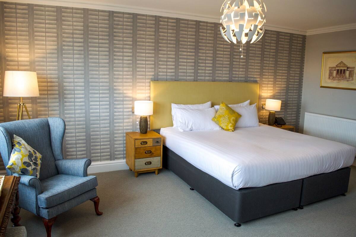 A hotel room with a large bed, two bedside tables with lamps, a chair, and a patterned wall.