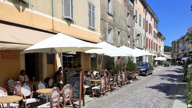 Un café a l'aire lliure amb caires i taules de vimet, sota ombrel·les blanques, situat en un carrer empedrat.