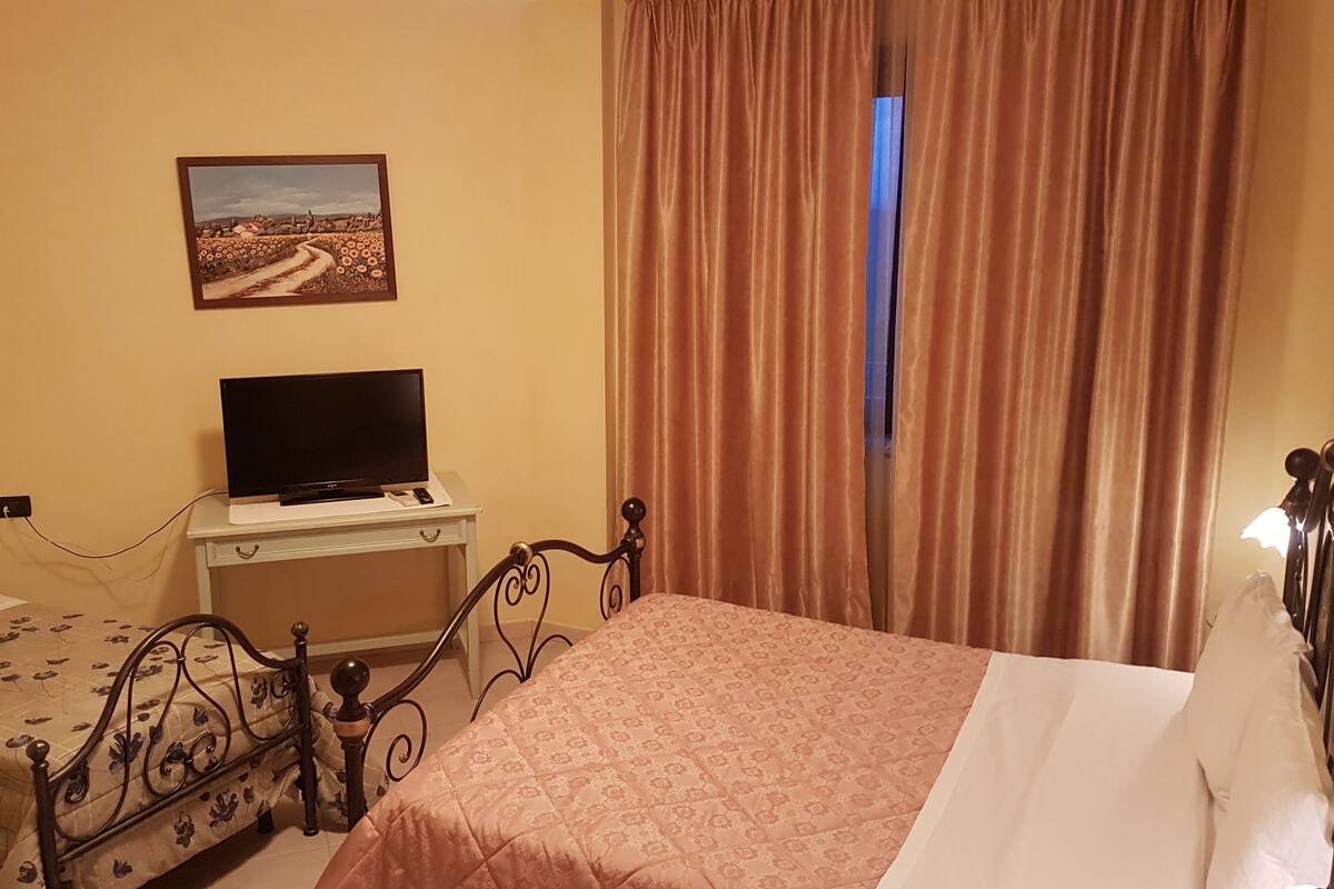 A hotel room with a bed, a TV on a bedside table, a painting on the wall, and a window with curtains.