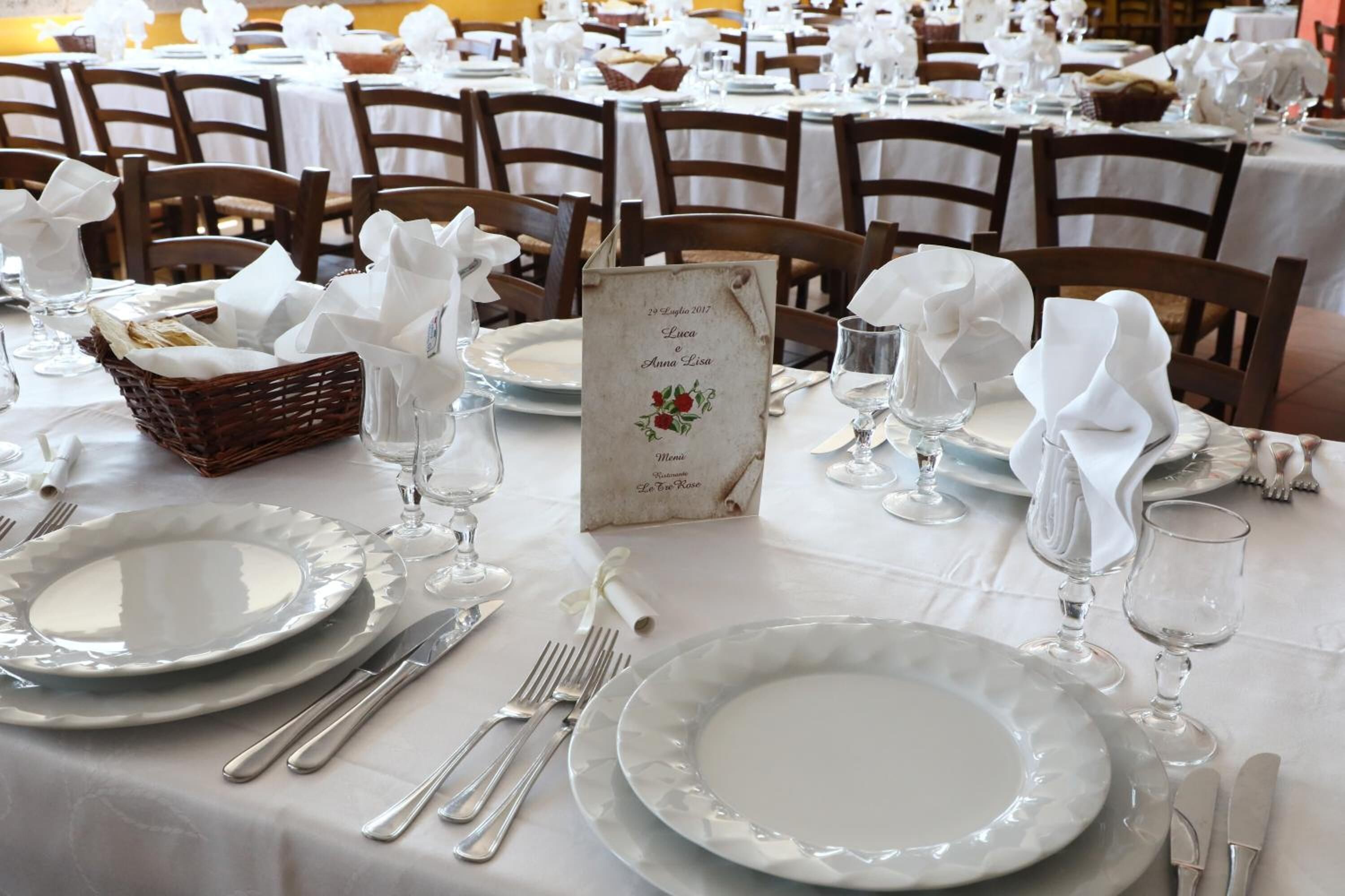 A dining table set with white plates, silverware, and glassware, with a menu stand and a basket of bread.