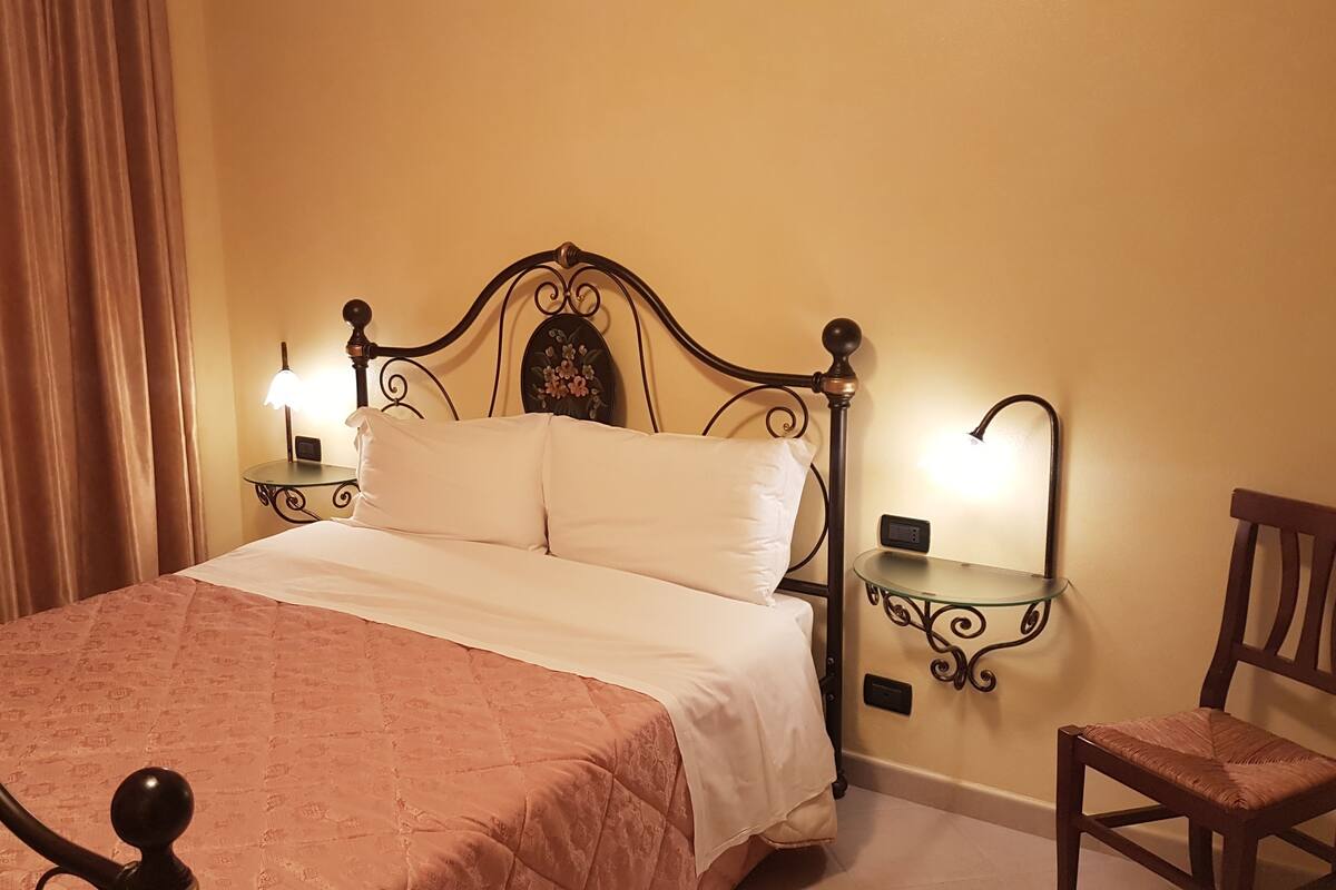 A neatly made bed with a headboard, two bedside tables with lamps, a chair, and a wall-mounted shelf.