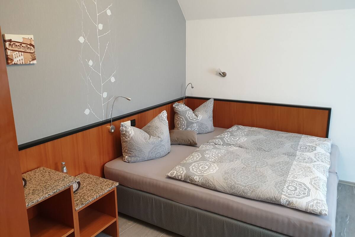 Een slaapkamer met een bed, twee kussens, een nachtkastje, een wandlamp en een klein plankje.