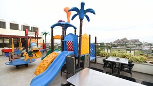 A playground with slides and climbing structures, a table, and chairs, overlooking a distant hill with a fort-like structure. - HOTEL ROCK FORT VIEW by Park Elanza (Tiruchirappalli)