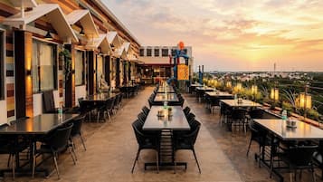 A rooftop restaurant with multiple tables and chairs, a view of the cityscape, and a sunset in the background.