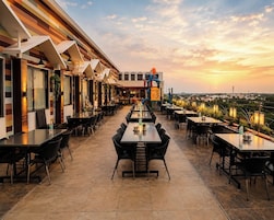 Un restaurante en la azotea con varias mesas y sillas, vistas panorámicas de la ciudad y una puesta de sol al fondo.