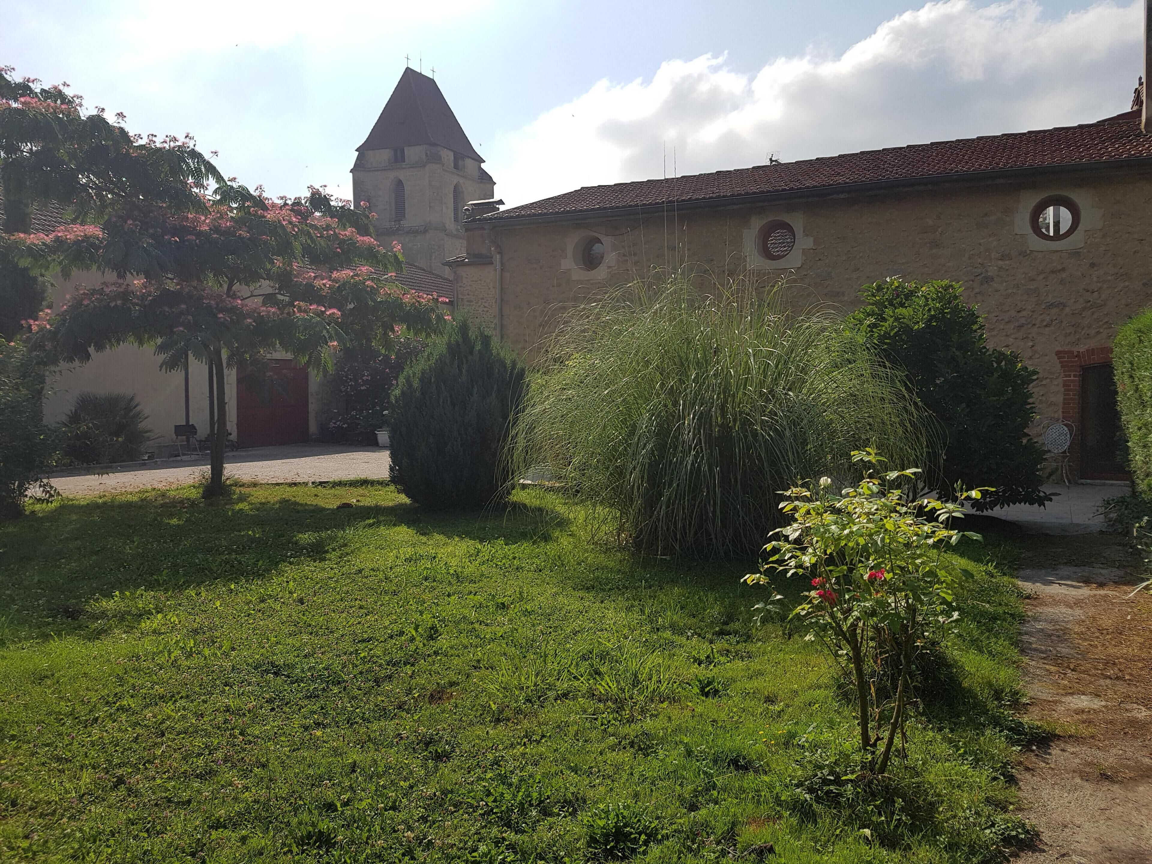 Un pati amb una torre d'església, un edifici de maó amb finestres circulars i vegetació abundant.