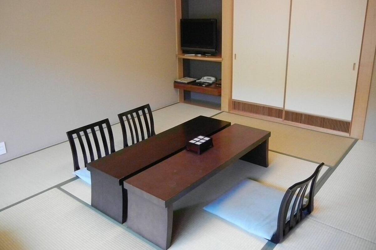 A traditional Japanese-style room with tatami flooring, a low table, four chairs, a television on a shelf, and sliding doors.