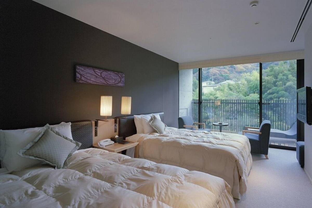 A hotel room with two beds, a nightstand, a desk, and a balcony with a view of trees.