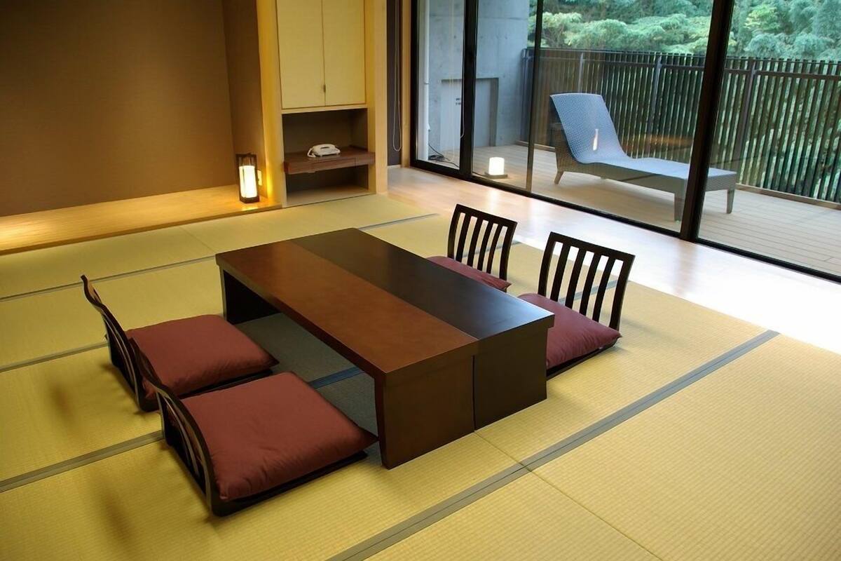 A tatami mat room with a low table, chairs, and a sliding glass door leading to an outdoor balcony with a chair.