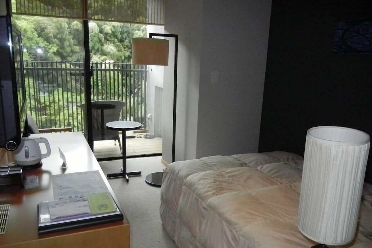 A hotel room with a bed, a desk with a lamp, a chair, and a view of greenery outside.