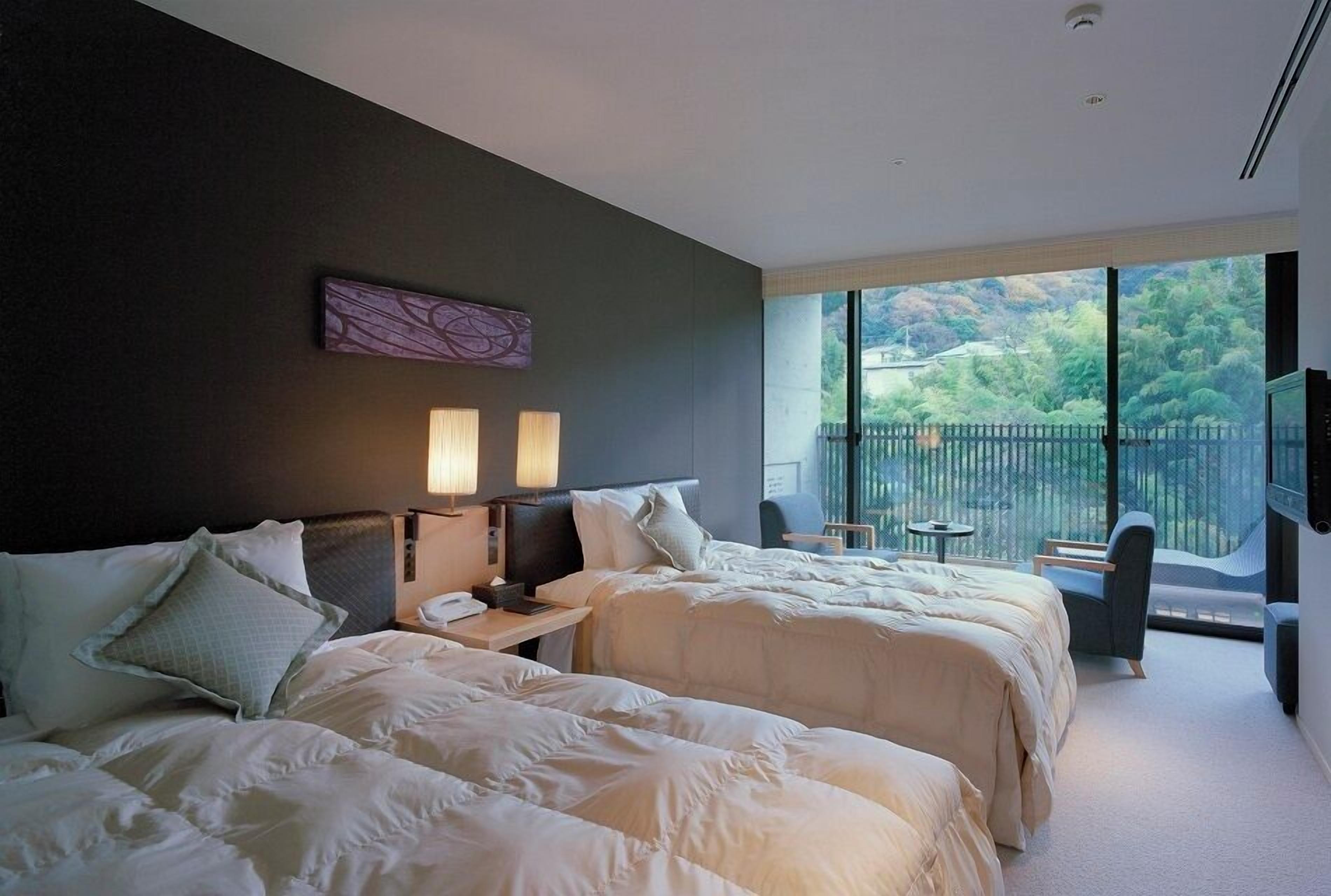 Habitación de hotel con dos camas, una mesita de noche, un escritorio, una silla y un balcón con vistas a la vegetación.