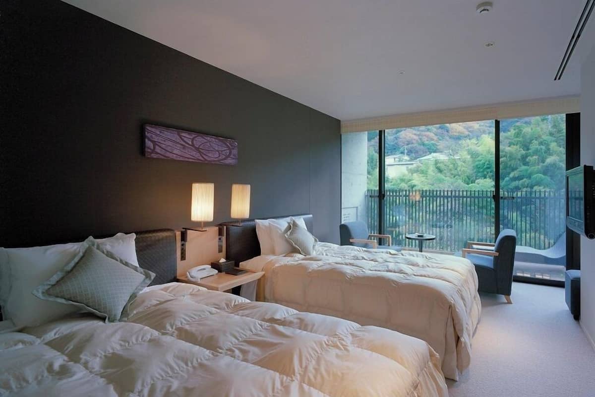 A hotel room with two beds, a nightstand, a desk, a chair, and a balcony with a view of greenery.